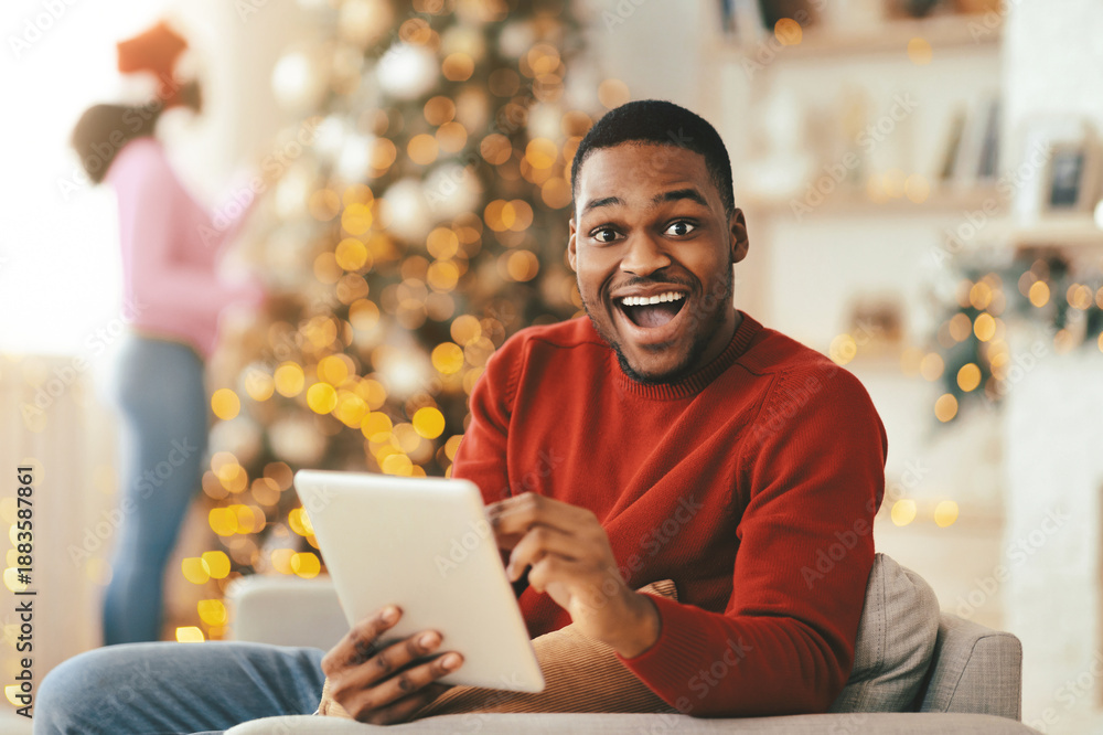 Obraz premium A man sits on a couch, looking happy while using a tablet. The living room is decorated for the holiday season with a Christmas tree and warm lights in the background.