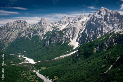 Panoramic summer view of the dramatic limestone peaks of the Accursed Mountains rising steeply above the lush green forests and dry riverbed of Valbona Valley in northern Albania
