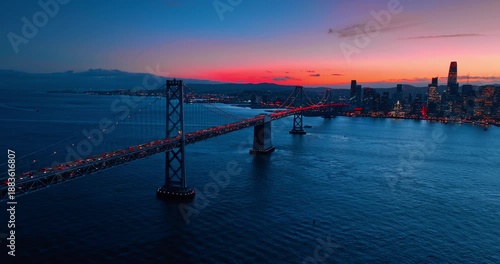 Cars with sparkling headlights move by the double-level bridge. Drone flight over the bay at dusk time. Orange sky at backdrop.