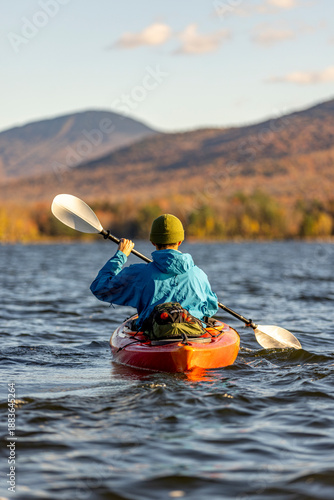 African-American man kayaking on a lake during peak fall autumn foliage with mountains and forest and view towards a ski area. Chittenden Reservoir, Vermont, New England, USA.