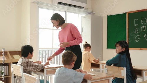 Caucasian teacher walking and check student homework at classroom while diverse children doing classwork at classroom. Diverse smart student lecturing and taking a note at elementary school. Pedagogy.
