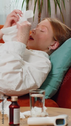 Teenager ill red hair lady suffering from cold or allergy lying on sofa couch in living room at home. Sick child blows sneezes wipes snot into napkin. Coronavirus quarantine, pandemic, flu. Lifestyles