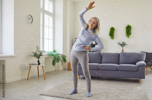 Happy senior woman doing physical exercise, side bends and stretching workout at home. Active old person training, grandmother standing on mat in living room with focus on mobility and balance posture