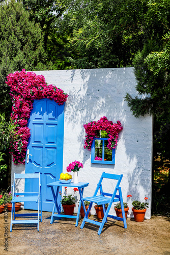 The photograph shows a dummy house with white walls, chairs and an entrance door with a blue window and beautiful red flowers hanging down.