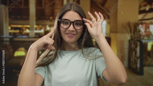 Teenager girl wearing casual top adjusts black eyeglasses in building corridor by hotel reception; confidence.