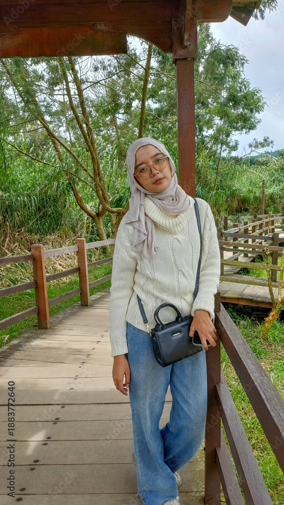 Obraz premium Young Asian woman in hijab and white sweater posing on wooden bridge at park. Fashion lifestyle in nature Indonesia. Beautiful girl with glasses. Sharp focus. Designer.