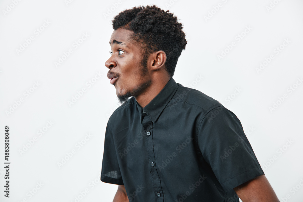 Fototapeta premium Young black man with dark skin and curly hair wearing black shirt looking surprised or amazed on white background. Side profile portrait of African ethnicity male with expressive face.