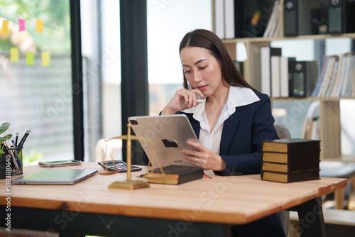 Asian businesswoman or lawyer in a formal blue suit sitting at a wooden desk, using a digital tablet. The desk is decorated with legal symbols including a gold scale of justice and law books. 