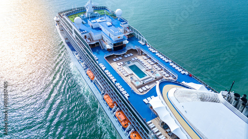 a luxury cruise ship's open deck featuring a swimming pool, jacuzzis, and rows of sunbeds. Multi-level recreational area with wooden flooring and blue decks over the ocean.