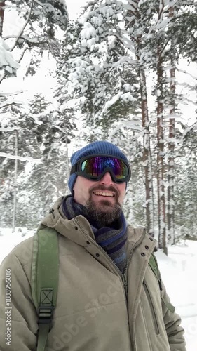 Winter explorer man standing in snowy pine forest, wearing insulated jacket, backpack, knit hat, scarf, mirrored ski goggles, surrounded by snow covered trees