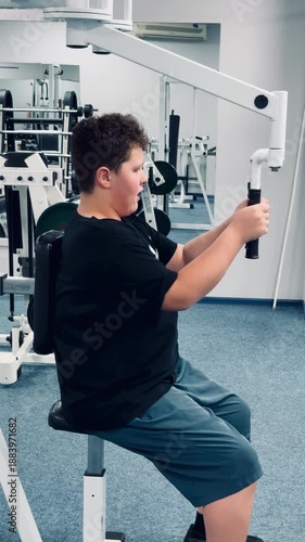 A boy is sitting on a bench in a gym. He is wearing a black shirt and blue shorts