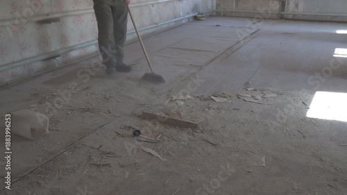 Man sweeping dust and debris on the floor of an empty, abandoned room using a broom or mop. The neglected space shows signs of decay and wear