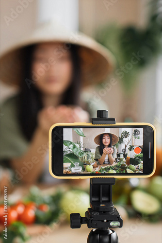 A young woman in a wide-brimmed hat records a video blog about healthy eating and fresh produce with her smartphone on a tripod