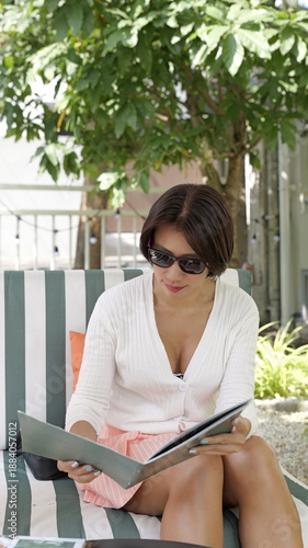 A stylish Asian woman in a white cardigan and pink skirt sits outdoors on a striped sofa while reviewing a menu