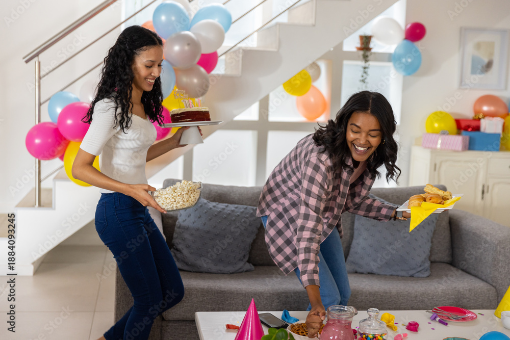 Obraz premium Diverse mom and teenage daughter carrying cake with candles and popcorn toward table