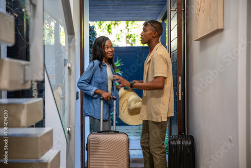 African American couple standing and talking at entryway with rose gold suitcase and straw hat