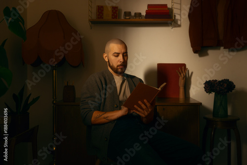 Slika na platnu A bearded man sits indoors reading a book in a softly lit, cozy room with warm tones and vintage details