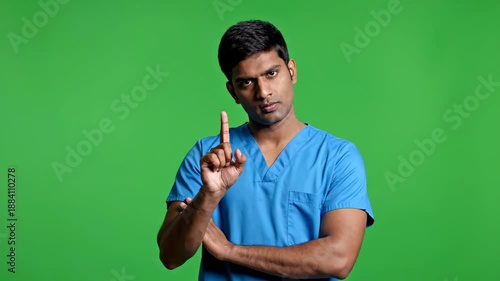 An Indian man in blue scrubs points upwards with a stern expression
