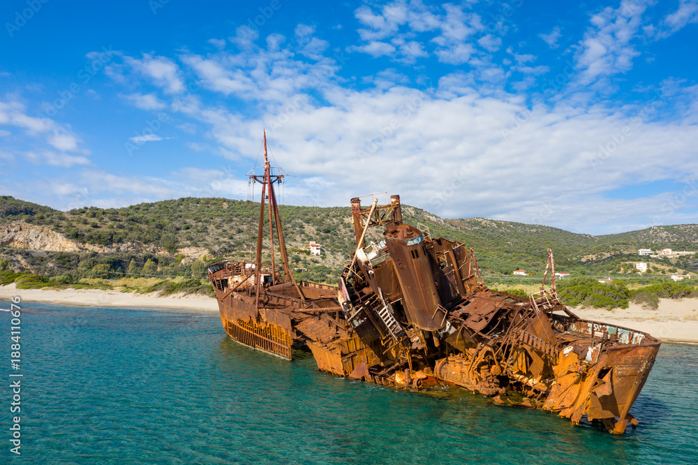 Fototapeta premium The Dimitrios shipwreck lies heavily rusted and broken in shallow turquoise water near a sandy beach, backed by green hills and scattered houses under a bright blue sky in Gythio, Greece.