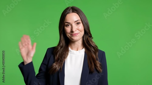 A woman smiles with a waving hand, wearing a blazer, with green background