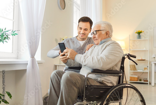 Fotografie Positive male nurse assisting an elderly patient in a wheelchair with a smartphone and online entertainment in internet at a nursing home