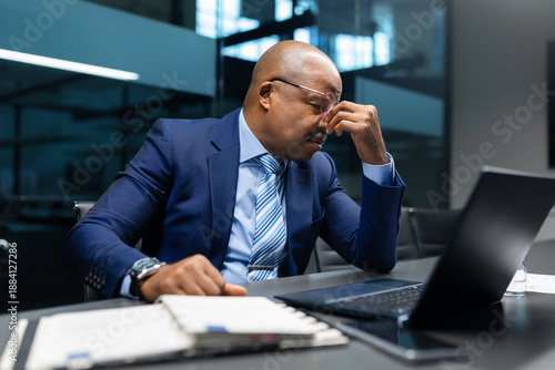 Tired black mature businessman rubbing his eyes at desk while working on laptop, illustrating exhaustion, stress, and high responsibility in executive corporate work