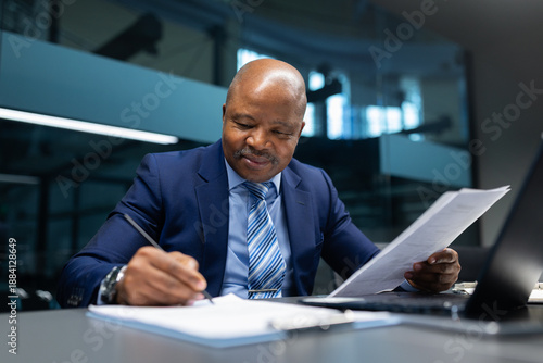 Confident African American businessman reviewing documents and signing paperwork at a modern office desk, focused professional workflow with laptop, pen, and corporate environment