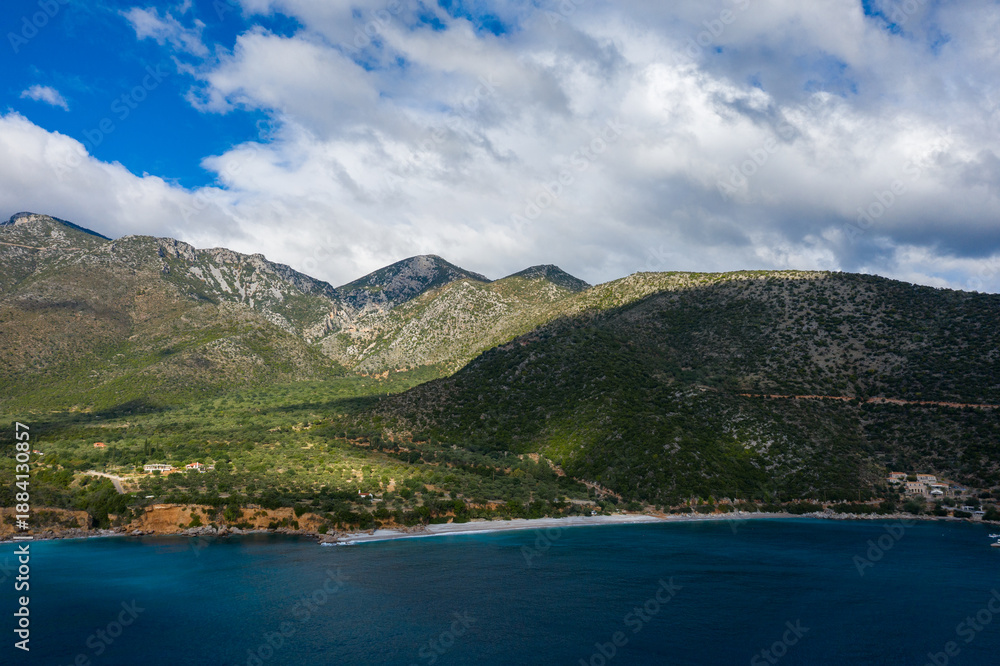Fototapeta premium A vibrant blue bay borders a secluded stretch of coastline, backed by lush, sun-dappled mountains under a sky of dramatic clouds. The landscape features rugged slopes, scattered houses, and a tranquil