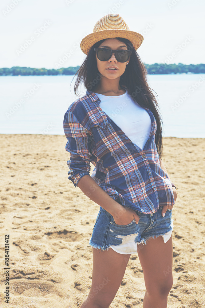 Fototapeta premium Woman, tourist and sunglasses at beach, outdoor and seaside with straw hat, pride and getaway in summer. Girl, trendy fashion and confident with shades, ocean or break in nature on island for holiday