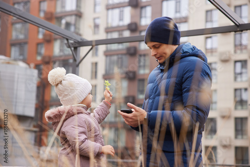 Young daughter presenting a green leaf to her father busy on smartphone in an urban setting