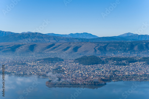 Foto A wide panoramic view captures the city of Ioannina and its island on Lake Ioannina, set against the rugged slopes of the Pindus mountains under a clear blue sky in Epirus, Greece