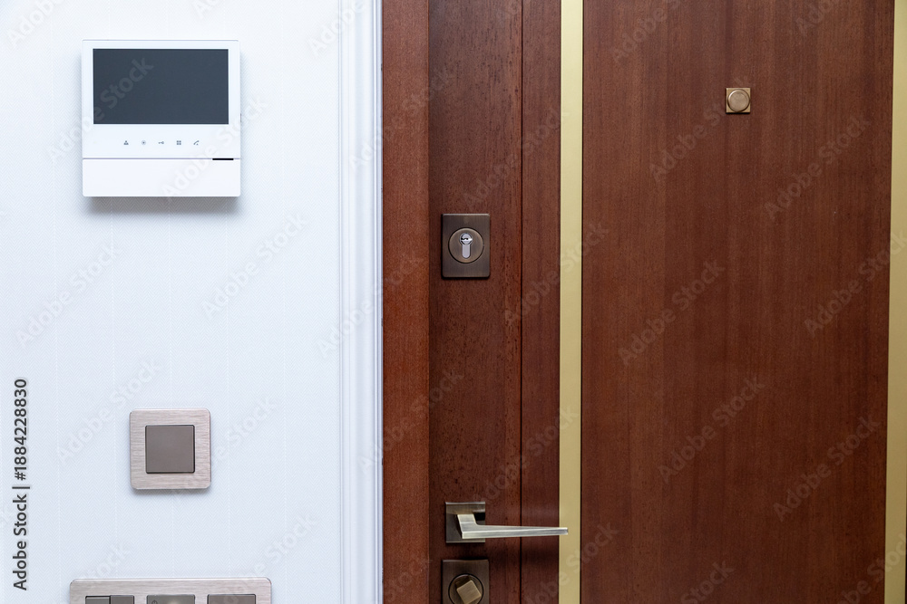 Obraz premium Modern wooden entrance door with gold fittings, peephole, intercom system on white wall and electrical switches, shows contemporary home design with functional elements for security and communication