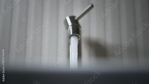 Close-up of minimalist metal faucet in modern bathroom. Human hand turns on and off tap and steady stream of clear water flows into sink. Soft light, gray textured wall, clean interior details
