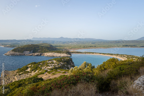 Wallpaper Mural Sweeping view of a crescent-shaped sandy bay bordered by rocky headlands and lush green vegetation, with calm turquoise water and distant mountains under a soft blue sky in Navarino Bay, Messenia. Torontodigital.ca
