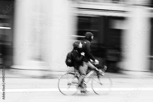 two young people riding a bicycle together in motion blur on a city street. Captured in black and white, the image conveys urban energy, youth and alternative transportation. Street photography.