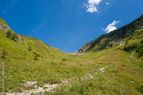 Wide view of a green alpine pass with rugged slopes and scattered rocks, bathed in bright sunlight beneath a vivid blue summer sky near Col du Tricot.