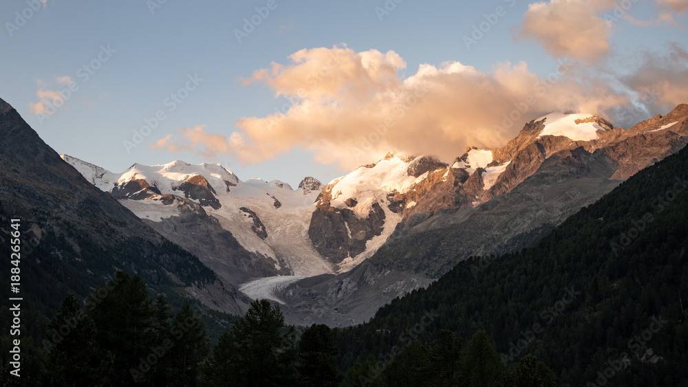 Obraz premium A breathtaking morning view captures the beauty of Piz Bernina, showcasing its glaciers and rugged peaks in the Swiss Alps, bathed in warm sunlight against soft clouds.