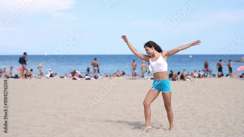 A woman stands on the beach in a blue bikini. She is smiling and she is enjoying herself. The beach is crowded with people, some of whom are also wearing bikinis. The atmosphere is lively and fun