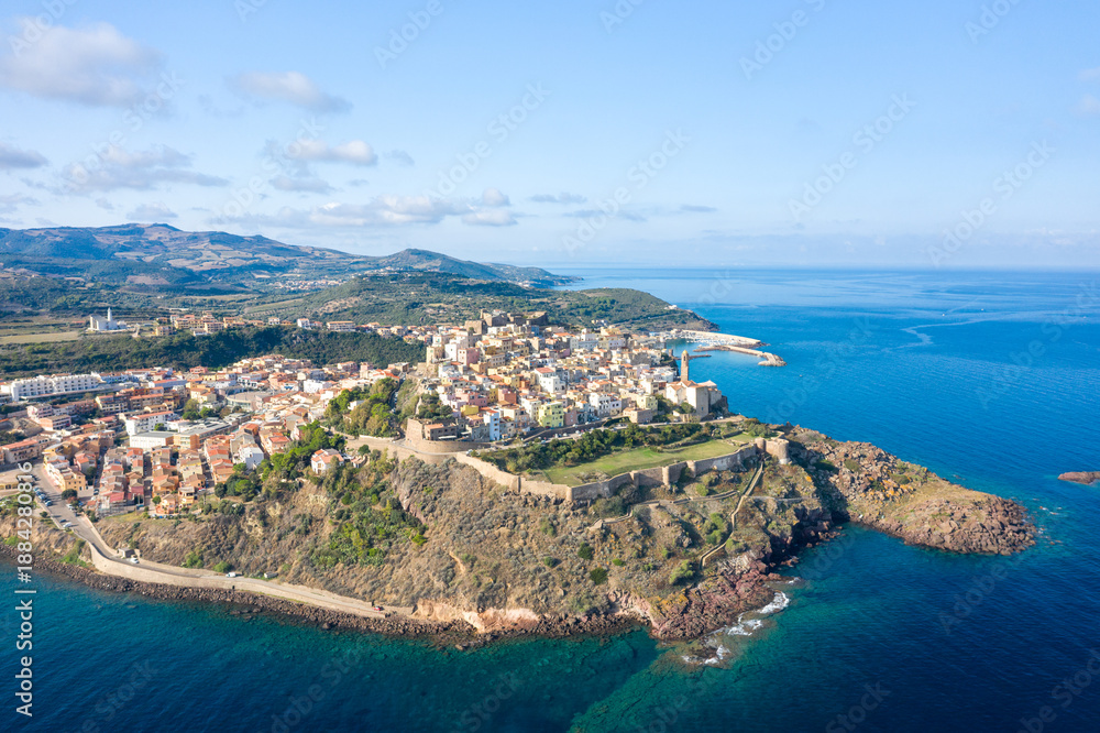 Fototapeta premium Sweeping aerial view of Castelsardo's historic center perched on rugged cliffs, with ancient fortress walls overlooking the vivid blue Mediterranean and rolling Sardinian hills.