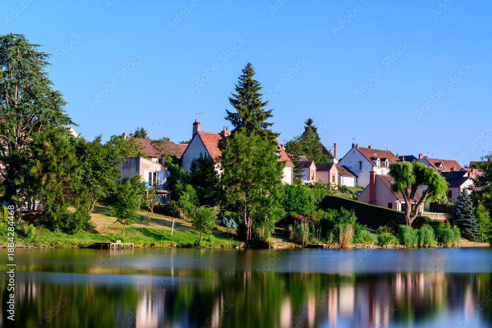 Obraz premium Quaint village homes with red-tiled roofs stand above a vibrant green shoreline, their forms reflected in the calm water of the pond beneath a bright blue sky. Summer sunlight and abundant foliage