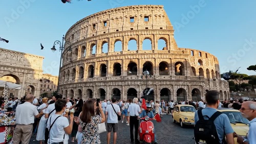 Colosseum view man gazes upon the ancient arena amid tourists, market stalls, and a red scooter in Rome, Italy