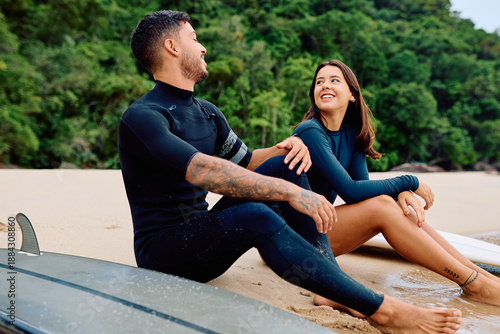 Young couple surfers in wetsuits sitting on sandy beach, smiling and having conversation after a surf session