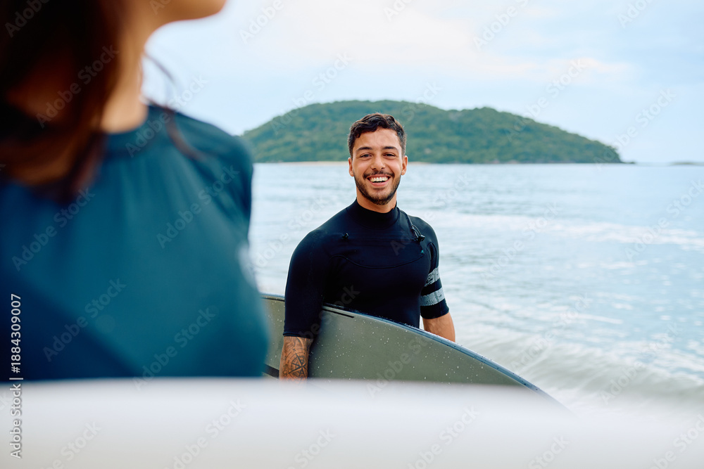 Fototapeta premium Young man wearing a wetsuit and holding a surfboard standing on the beach with the ocean and an island in the background