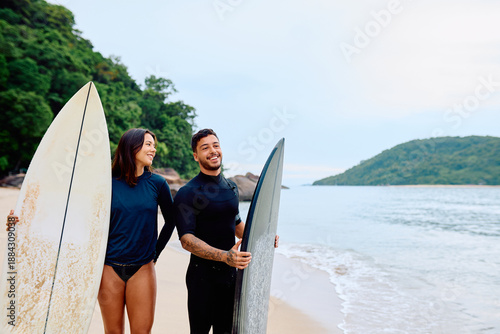 Canvas Print Young couple wearing wetsuits walking on a tropical beach with surfboards, antic