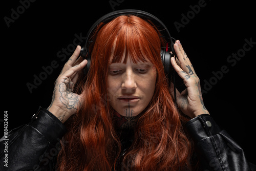 A DJ wears headphones and focuses on the music. Long red hair frames the face, while decorated hands adjust the headphones. The setting is simple with a dark background.