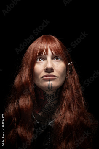 A DJ stands with red hair and tattoos, looking upwards in a dark environment. The setting highlights the person's expression and style during a music event.