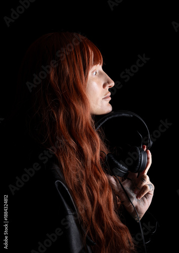 A DJ stands in a dark space, holding headphones while looking to the side. The long red hair contrasts with the background. The person appears to be focused on their music.