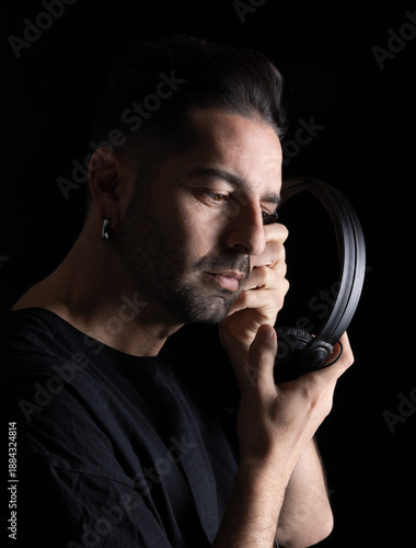 A DJ focuses while holding headphones. He is in a dark setting, engaged in music preparation. The quiet moment highlights his concentration and passion for his craft.
