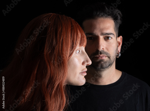 A couple stands closely together in a dark setting. The person on the left has long red hair and a unique look, while the person on the right is serious and focused. They are dressed in black.