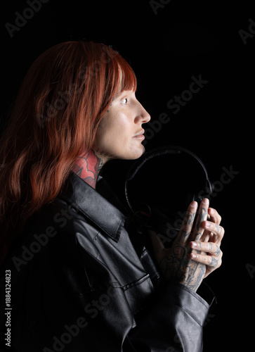 A woman DJ stands in a dark space, holding headphones close to her face. Her long red hair and tattoos are visible, highlighting her focus on the music and the moment.